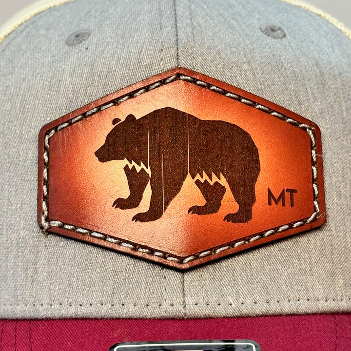 A close up of a made in Montana hand dyed leather patch of an etched Bear silhouette and MT, sewn on a Richardson trucker hat featuring a maroon bill, heather grey front and cream mesh.