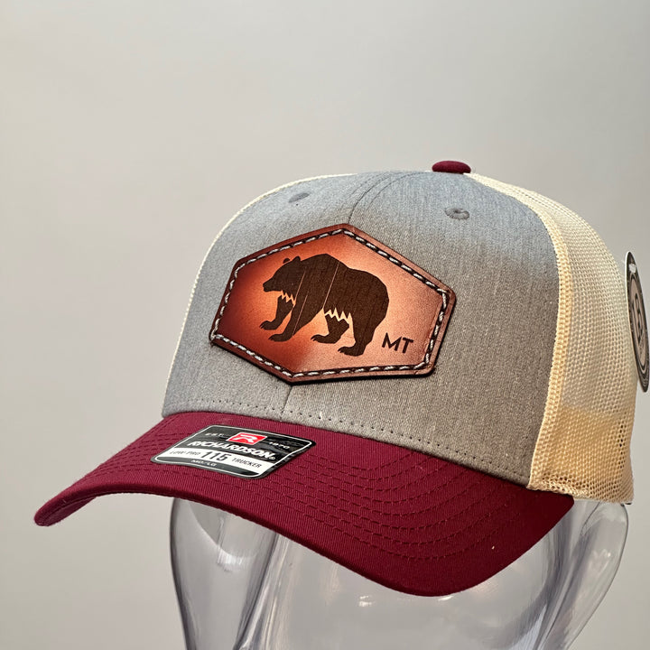 The side of a made in Montana hand dyed leather patch of an etched Bear silhouette and MT, sewn on a Richardson trucker hat featuring a maroon bill, heather grey front and cream mesh.