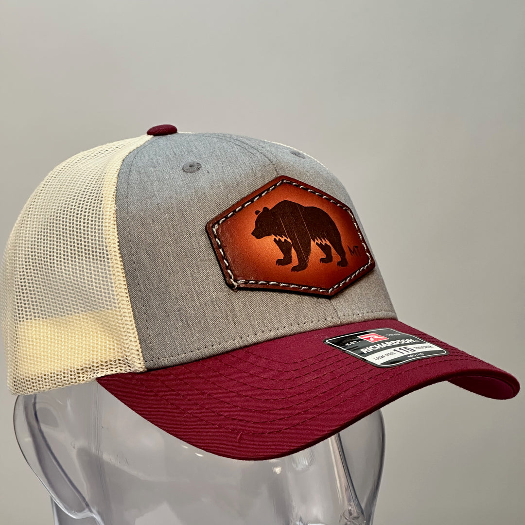 The side of a made in Montana hand dyed leather patch of an etched Bear silhouette and MT, sewn on a Richardson trucker hat featuring a maroon bill, heather grey front and cream mesh.