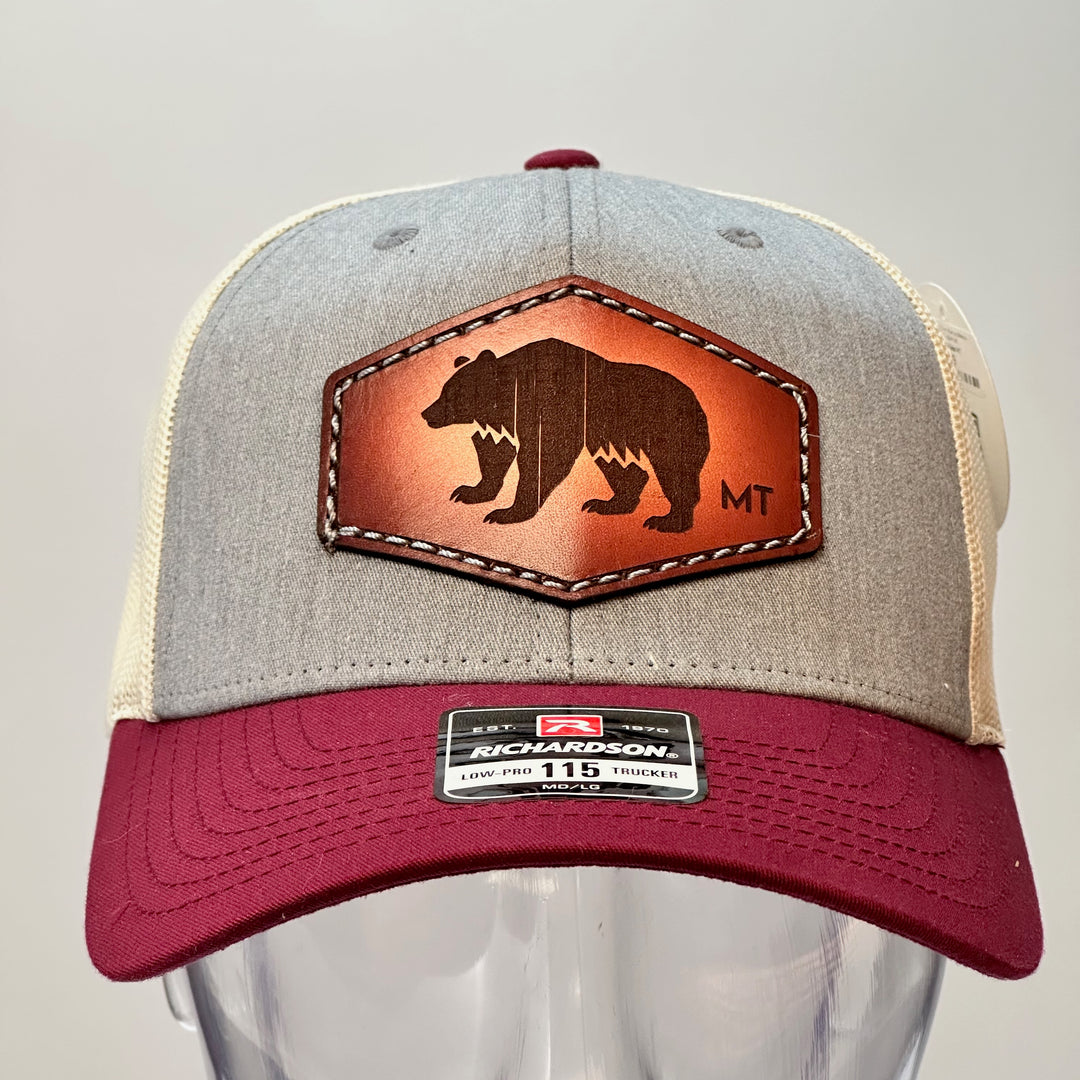 The front of a made in Montana hand dyed leather patch of an etched Bear silhouette and MT, sewn on a Richardson trucker hat featuring a maroon bill, heather grey front and cream mesh.