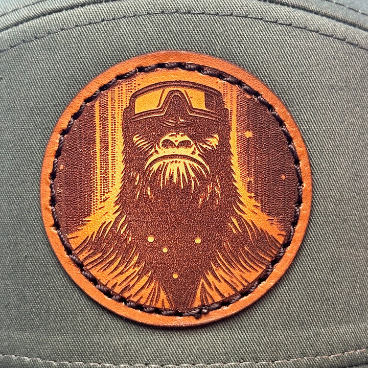 A close up of a hand dyed leather patch hat with sasquatch's face sewn onto a green Richardson flat bill trucker hat with black mesh.