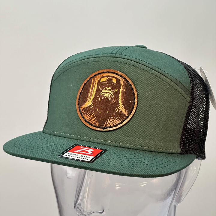 the side of a hand dyed leather patch hat with bigfoot's face sewn onto a green Richardson flat bill trucker hat with black mesh.