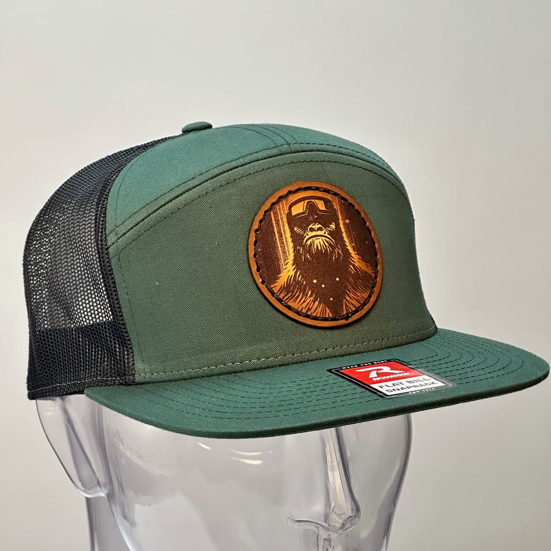 The side of a hand dyed leather patch hat with bigfoot's face sewn onto a green Richardson flat bill trucker hat with black mesh.