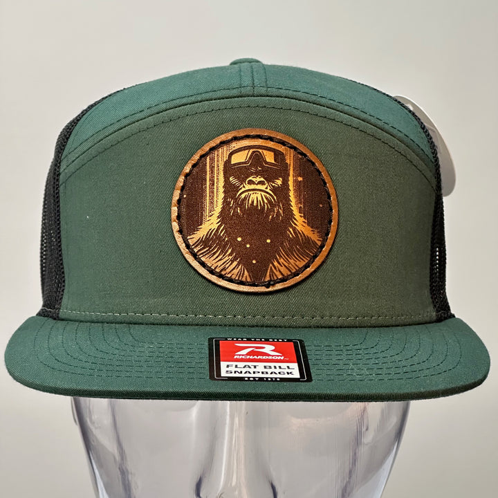 The front of a hand dyed leather patch hat with bigfoot's face sewn onto a green Richardson flat bill trucker hat with black mesh.