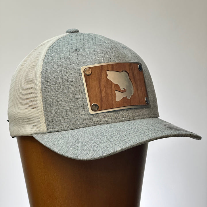 A side view of a cherry wood and metal patch with a walleye silhouette featured on a bright silver patch. Riveted to a heather grey and white flexfit trucker hat.