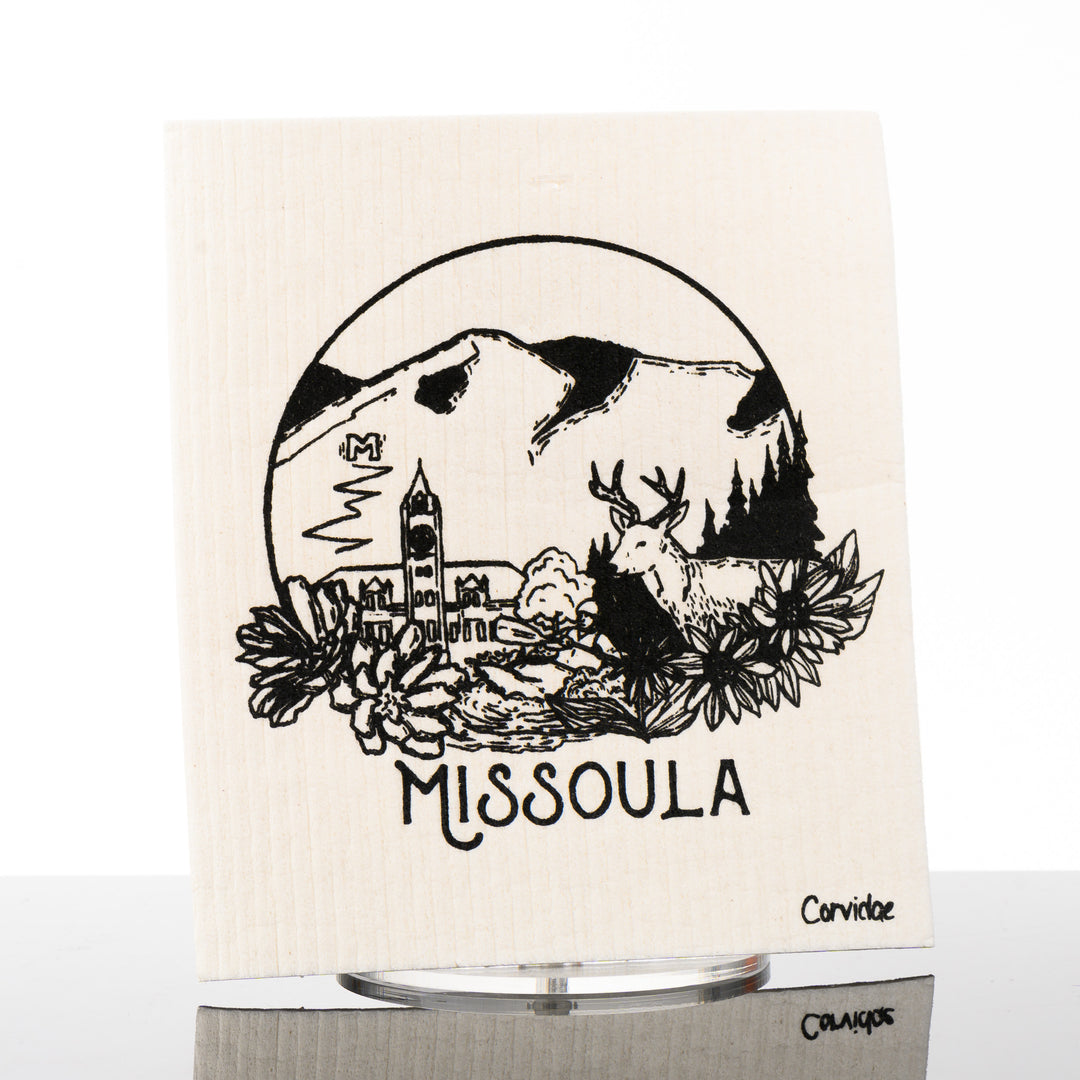 A rectangular swedish dish cloth with a drawing of Missoula with the M, the Clocktower, an elk and some bitterroot flowers.