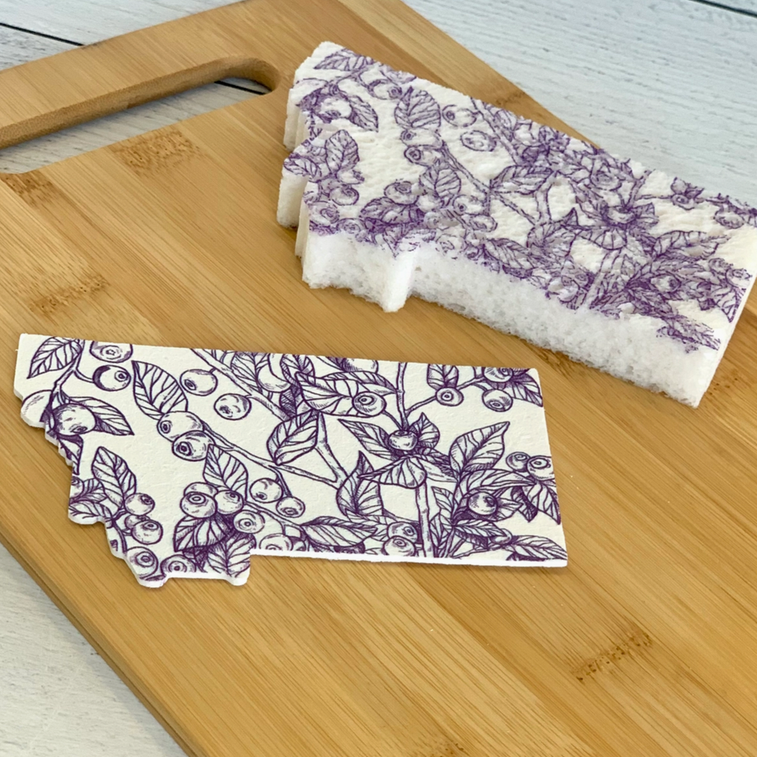 This Montana shaped pop-up sponge features a huckleberry hand drawn print. This image shows the difference between the sponge after it has popped up versus before. It has absorbed water and is flat.