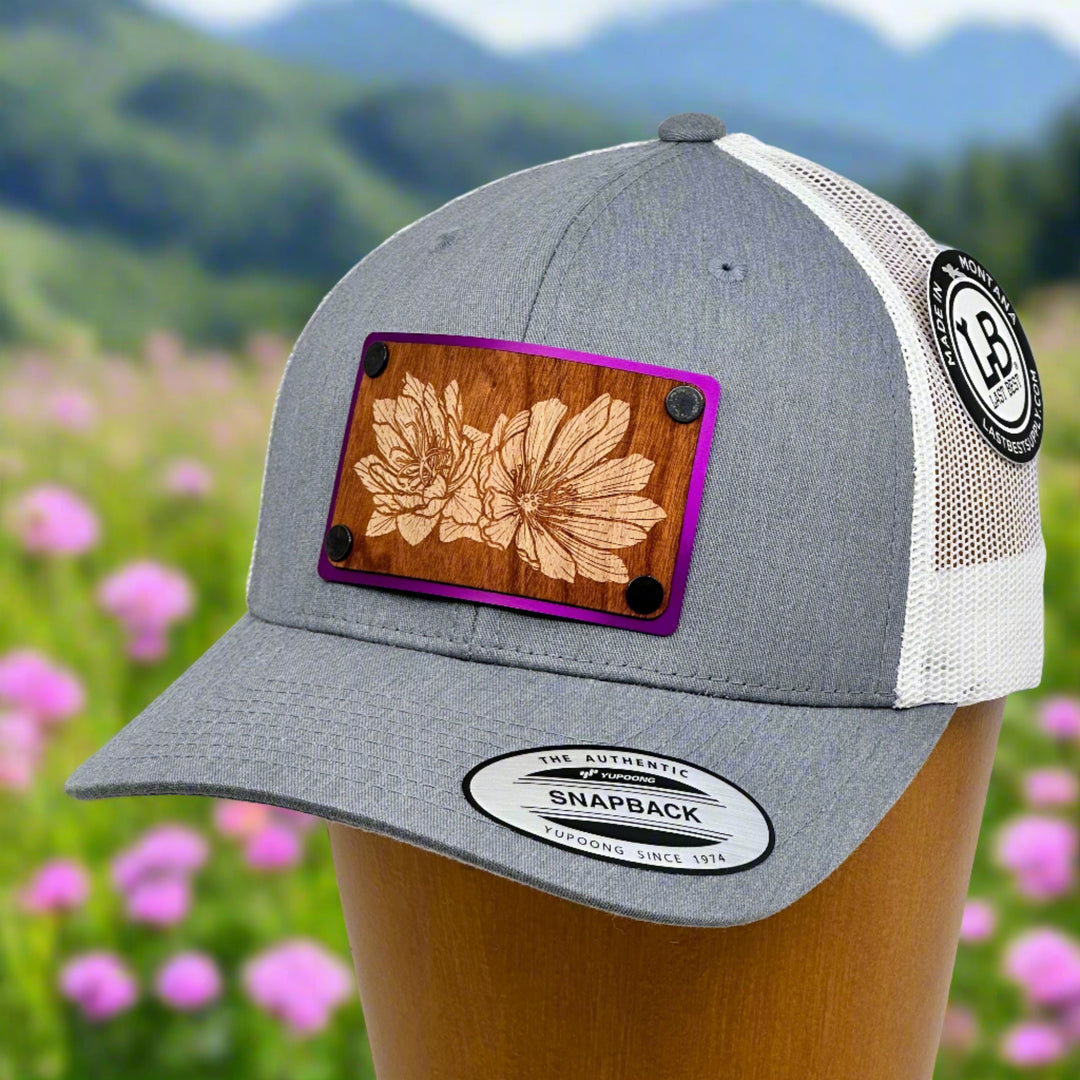 A grey and white trucker hat with an etched bitterroot art cherry wood patch  with a meadow and mountains in the distance.