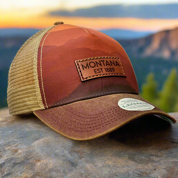 A 3/4 view of a trucker hat printed with mountains and khaki mesh and a thick leather patch that reads Montana EST 1889