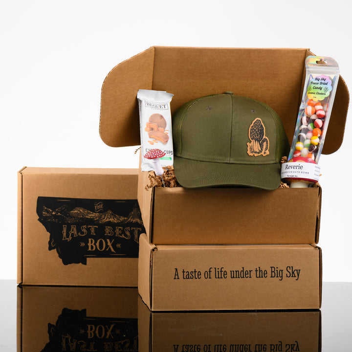 A stack of Last Best Boxes with a green hat with a morel mushroom wood patch, a mushroom bath bomb, freeze dried skittles, a mushroom plant decoration, and caramel chocolate cups.