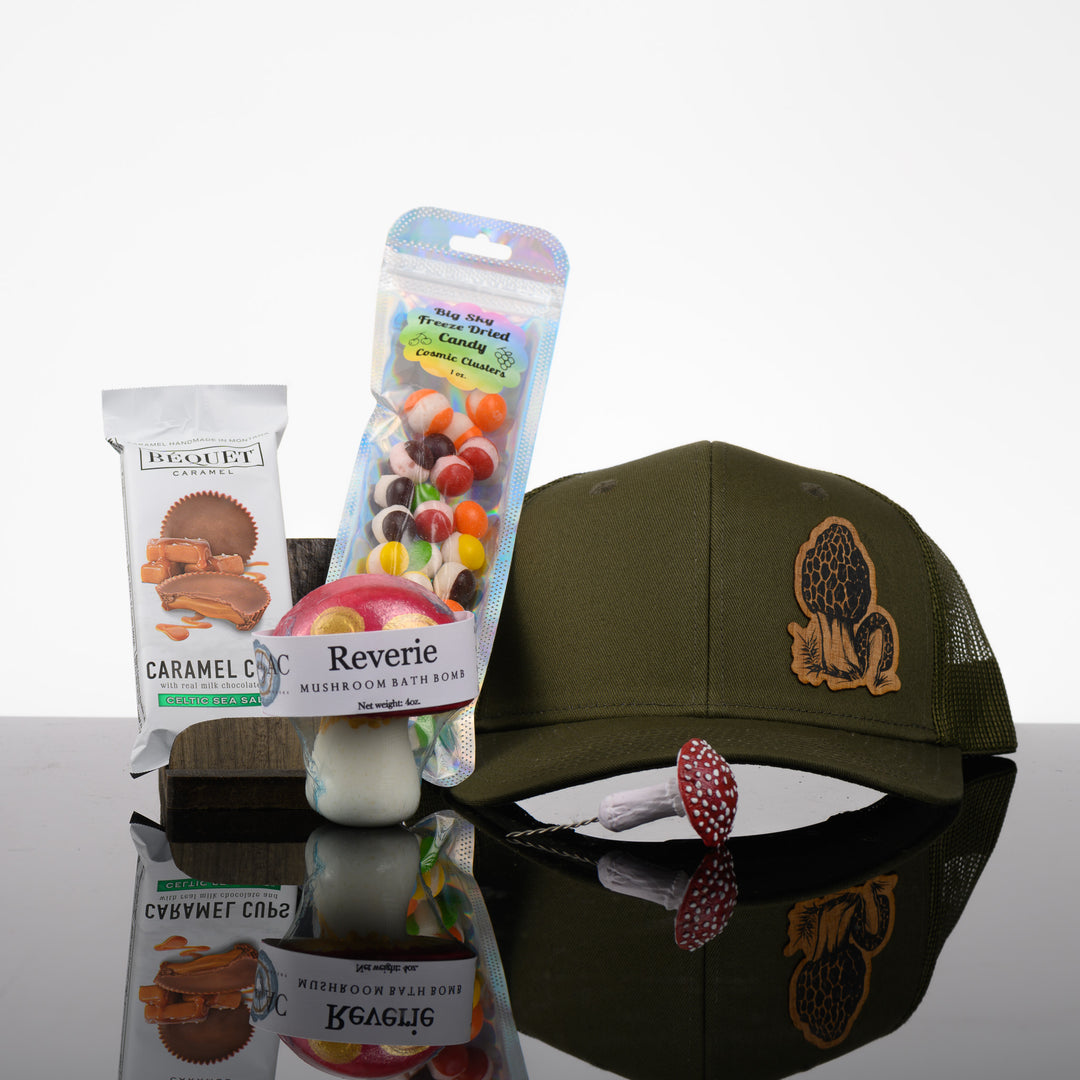 A group shot of the items in this box including green hat with a morel mushroom wood patch, a mushroom bath bomb, freeze dried skittles, a mushroom plant decoration, and caramel chocolate cups.