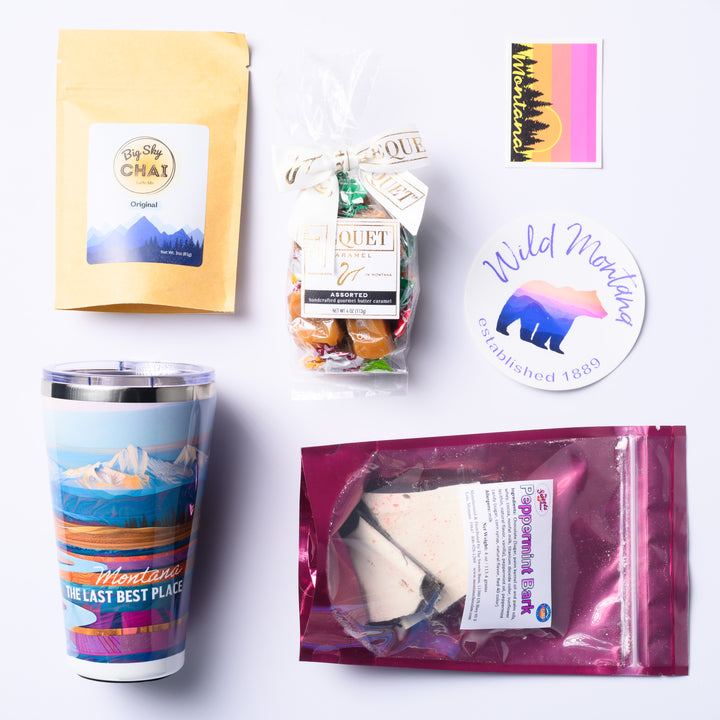 A flat lay picture of the Last Best Box, a Montana made gift box featuring Big Sky Chai, Peppermint Bark, Stickers, Bequet Caramel and a Montana scenery Tumbler.