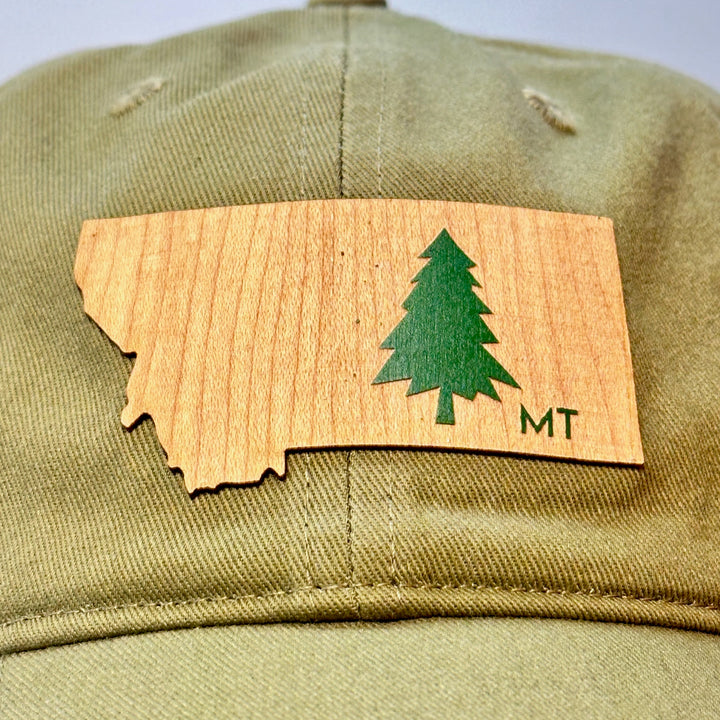 A made in Montana flexwood patch in the shape of Montana with an evergreen tree and the letters MT heat pressed onto an all cloth hat