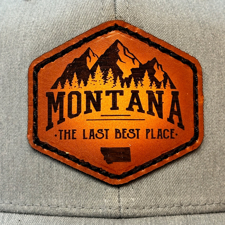 A close up view of a Made in Montana leather patch that says Montana, The Last Best Place and is sewn to a heather Grey and black trucker hat.