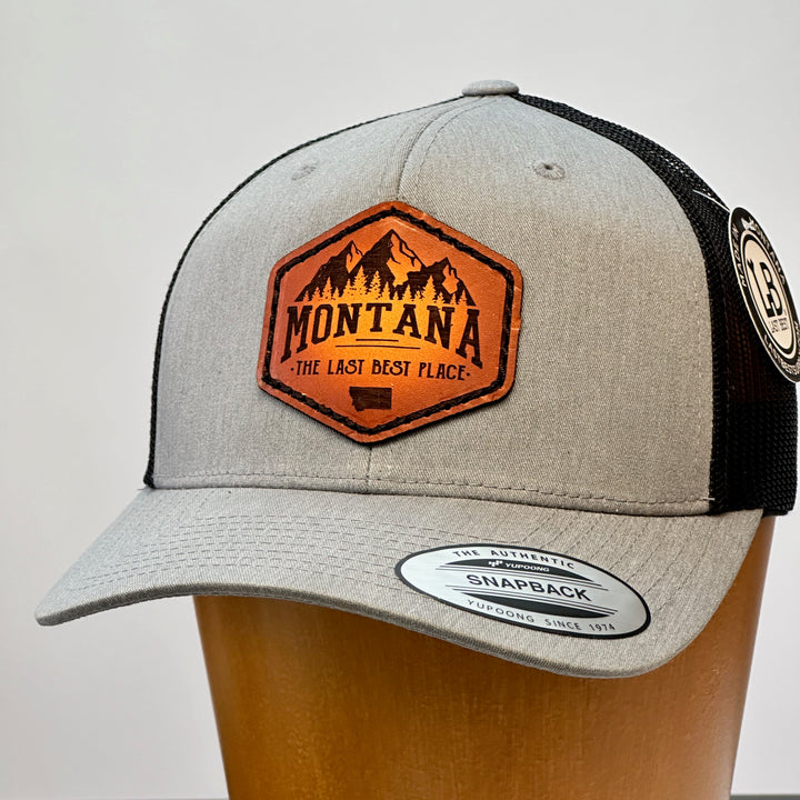 A side view of a Made in Montana leather patch that says Montana, The Last Best Place and is sewn to a heather Grey and black trucker hat.