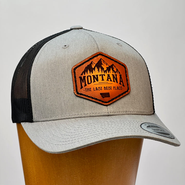 A side view of a Made in Montana leather patch that says Montana, The Last Best Place and is sewn to a heather Grey and black trucker hat.