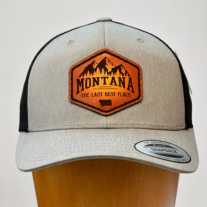 A Made in Montana leather patch that says Montana, The Last Best Place and is sewn to a heather Grey and black trucker hat.