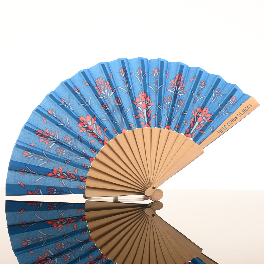 A Montana made artist hand fan featuring a blue background with hand drawn Indian Paintbrush flowers.