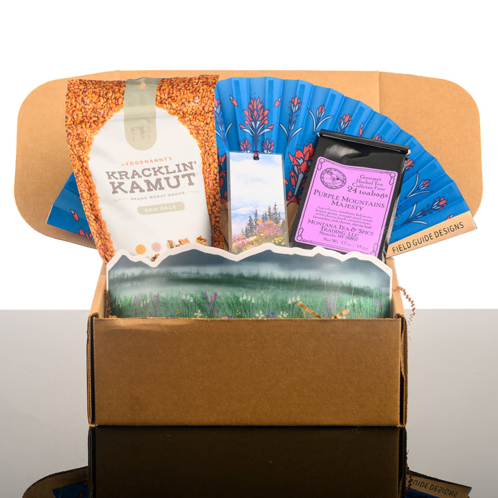 A Montana made gift box featuring a bag of kamut, a hand fan, a watercolor bookmark, purple mountain magesty tea and a large sticker wrap. 