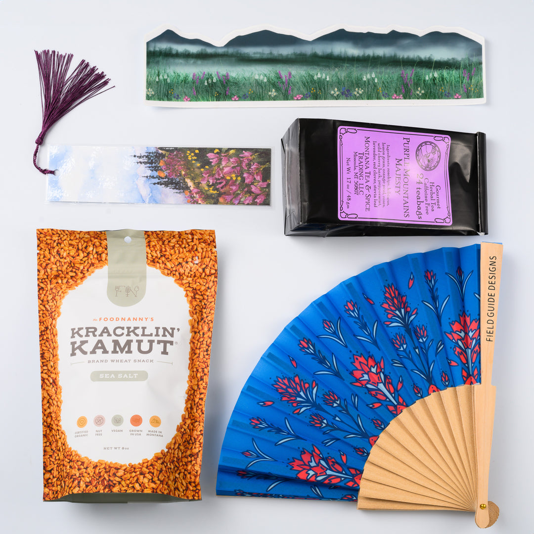 A flat lay image of a Montana made gift box featuring a bag of kamut, a hand fan, a watercolor bookmark, purple mountain magesty tea and a large sticker wrap. 