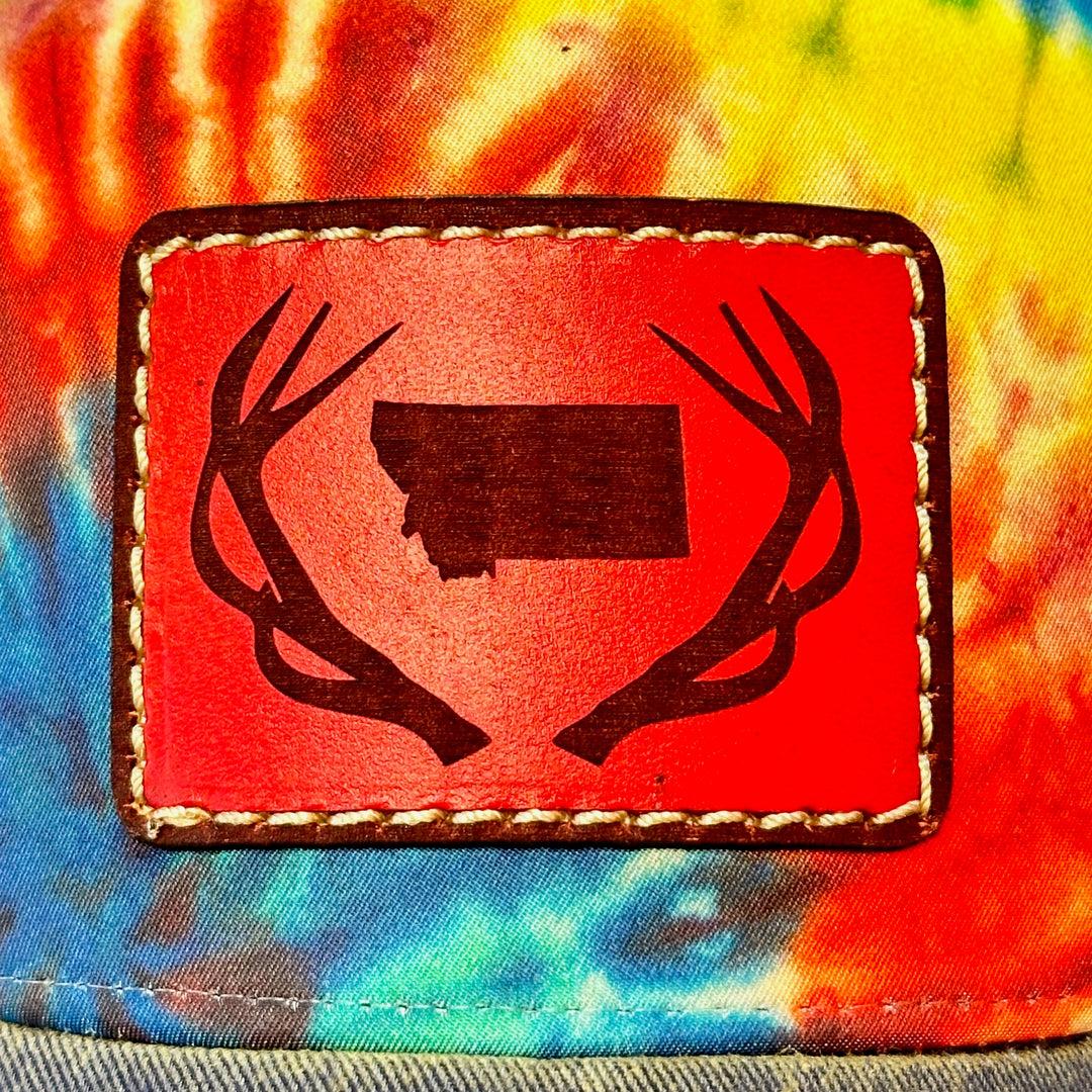 A close up of a hand dyed leather patch, made in Montana, of he shape of Montana with antlers around it, sewn to a rainbow tie dye Legacy trucker hat.