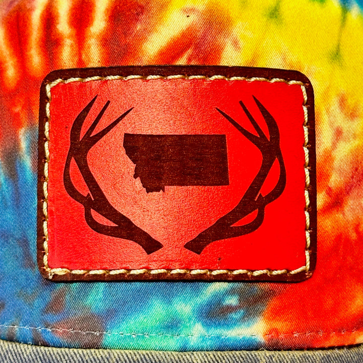 A close up of a hand dyed leather patch, made in Montana, of he shape of Montana with antlers around it, sewn to a rainbow tie dye Legacy trucker hat.