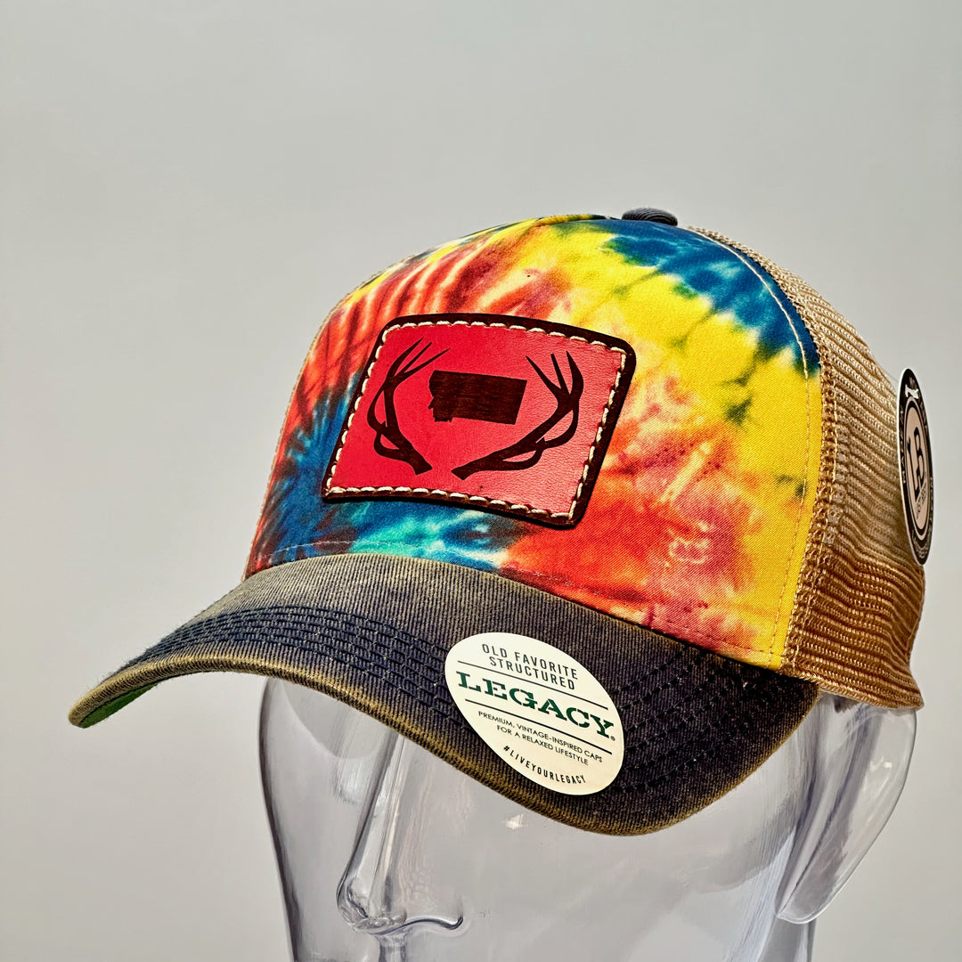 The side of a hand dyed leather patch, made in Montana, of he shape of Montana with antlers around it, sewn to a rainbow tie dye Legacy trucker hat.