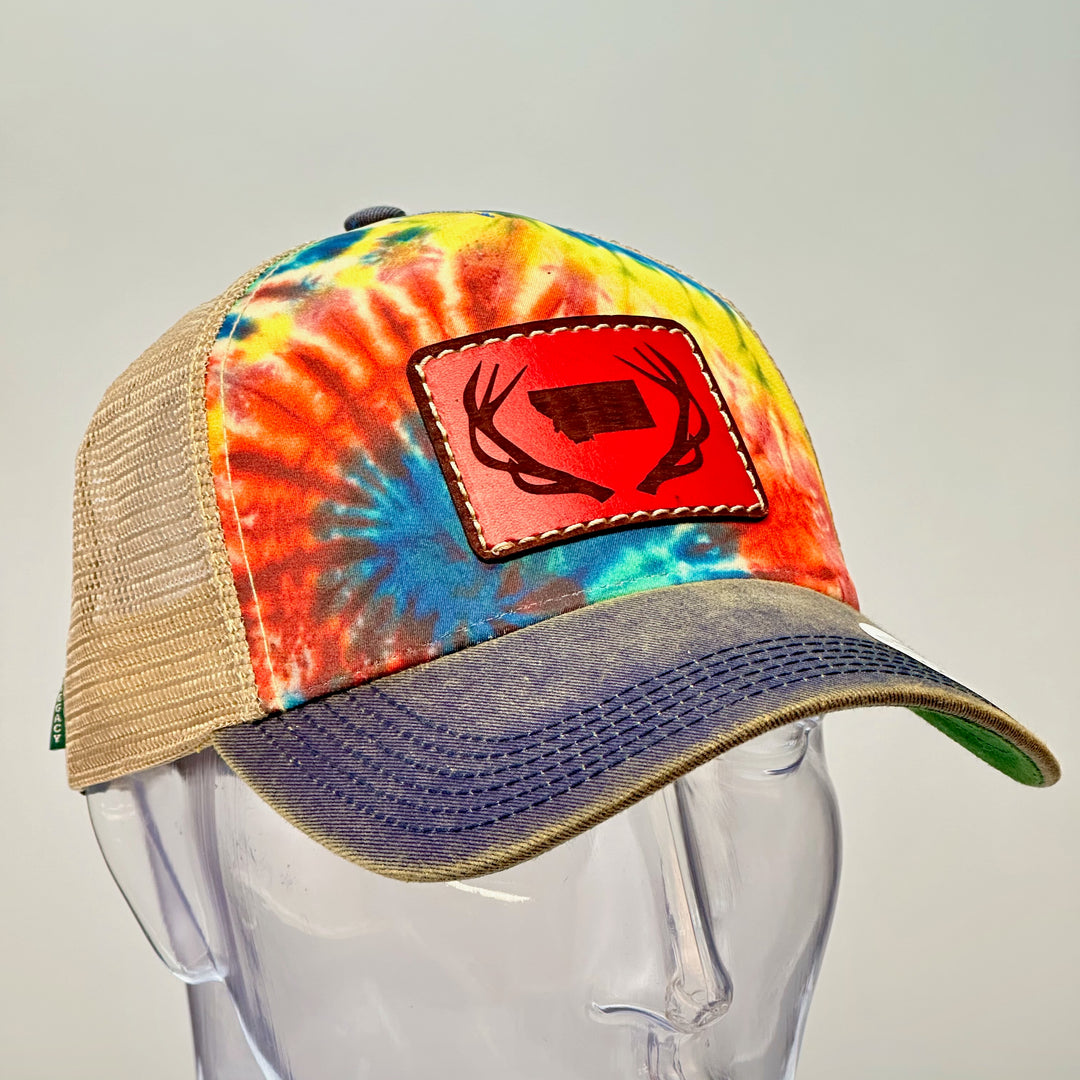 A hand dyed leather patch, made in Montana, of he shape of Montana with antlers around it, sewn to a rainbow tie dye Legacy trucker hat.