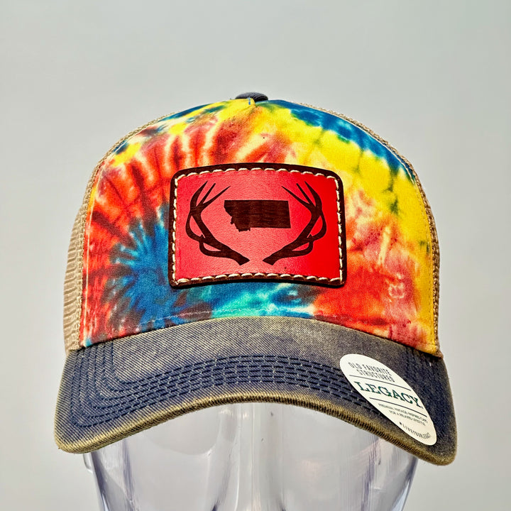 A hand dyed leather patch, made in Montana, of he shape of Montana with antlers around it, sewn to a rainbow tie dye Legacy trucker hat.