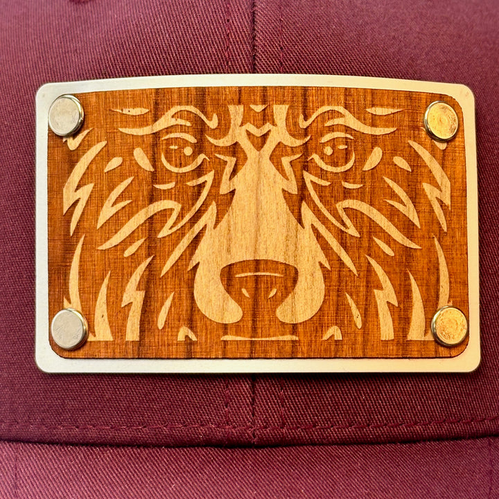 A close up view of a made in Montana bear face cherry wood patch maroon and grey trucker hat.