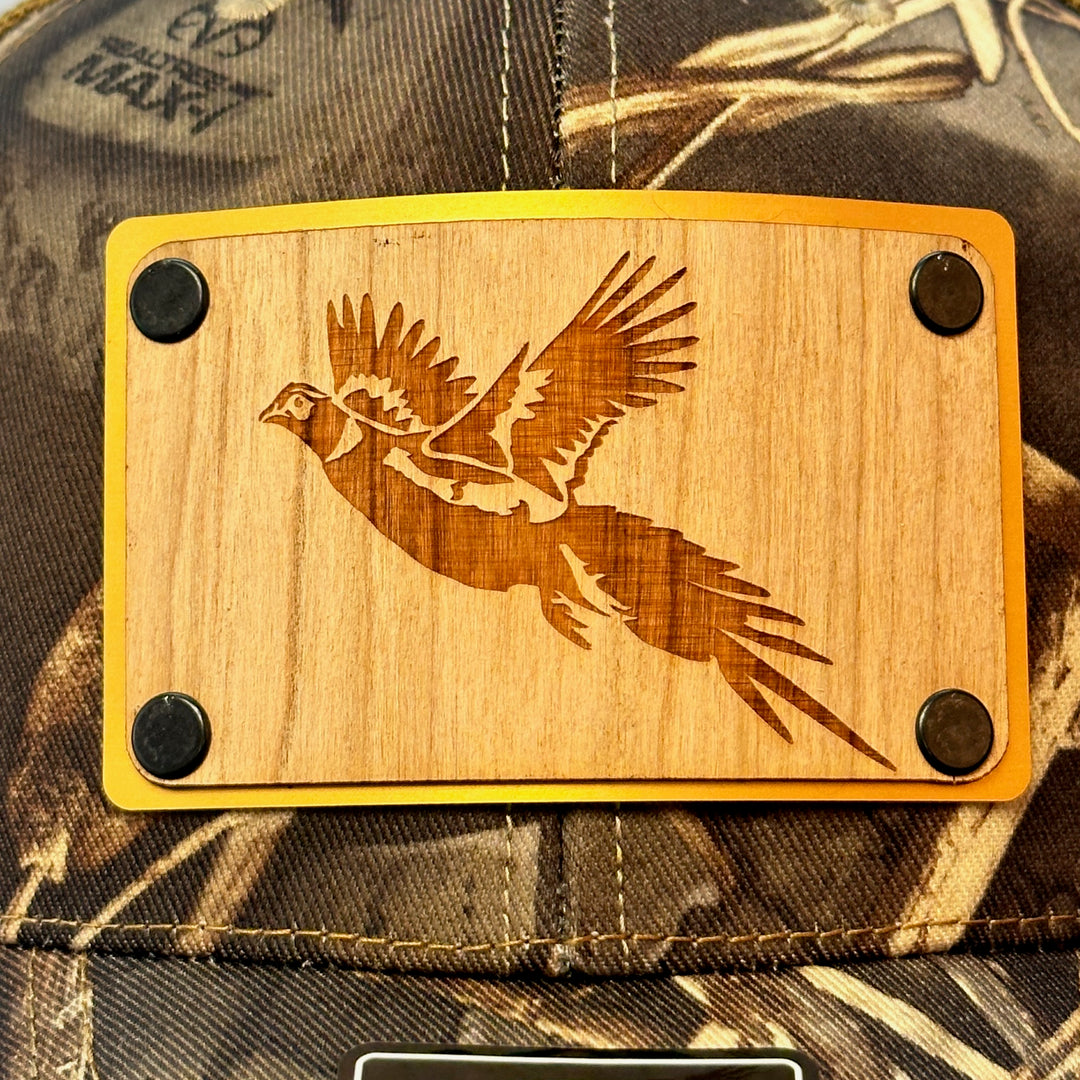 A close up of a made in Montana wood patch of an etched pheasant on a gold metal plate riveted with black rivets to a real tree camo Richardson trucker hat.