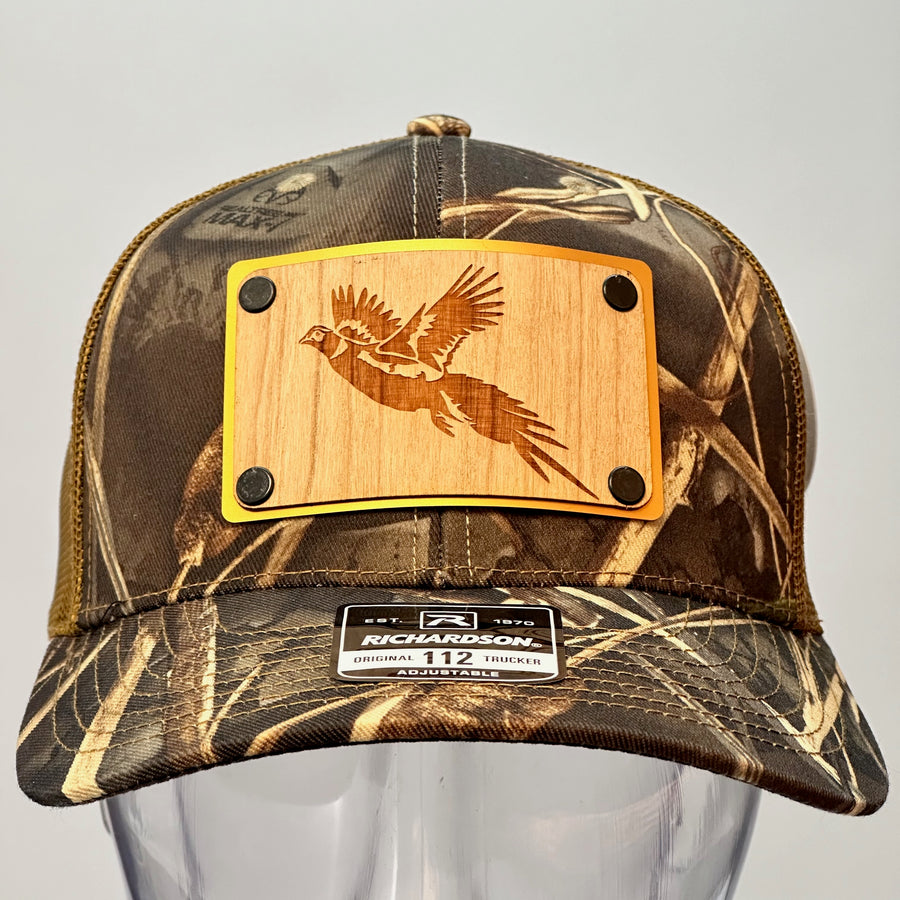 A front view of a made in Montana wood patch of an etched pheasant on a gold metal plate riveted with black rivets to a real tree camo Richardson trucker hat.